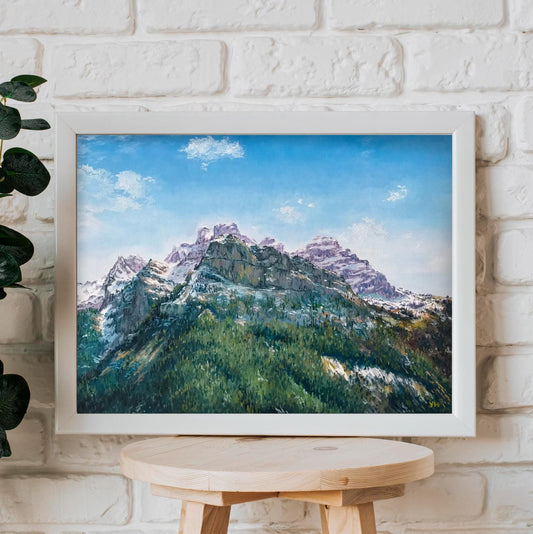Framed landscape print of mountains on a white brick wall with a plant and wooden stool.